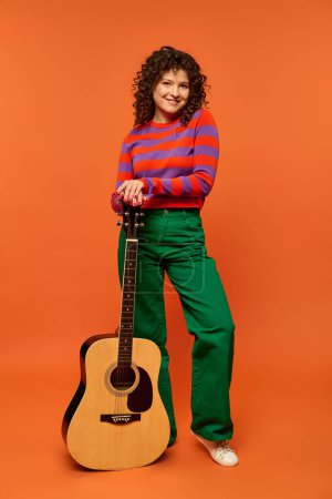 A young woman with curly hair poses with a guitar in front of a vibrant orange backdrop. She wears a striped sweater and green pants.