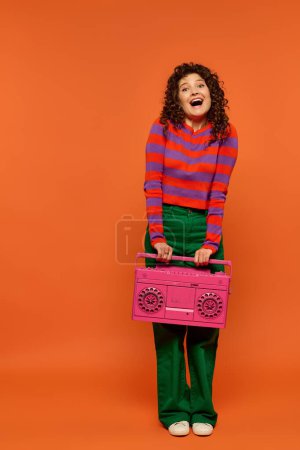 Eine Frau mit lockigem Haar hält eine pinkfarbene Boombox vor orangefarbenem Hintergrund und trägt ein gestreiftes Hemd, eine grüne Hose und weiße Schuhe..