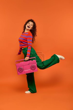 Eine junge Frau mit lockigem Haar in lebendiger Kleidung posiert vor leuchtend orangefarbenem Hintergrund und hält eine pinkfarbene Boombox in der Hand..