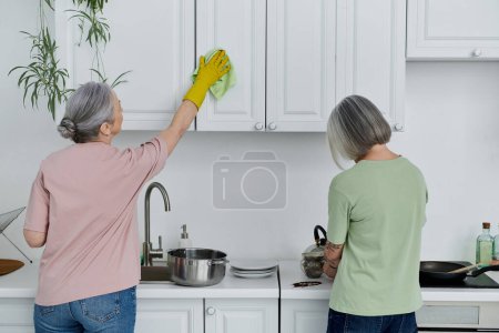 A lesbian couple cleans their modern kitchen together, one woman wiping cabinets while the other stands at the counter.