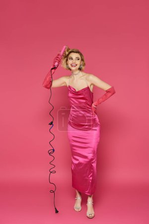A woman in a pink dress and gloves styles her hair with a curling iron.