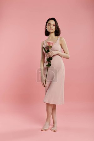 A woman in a pink dress poses with a rose in front of a pink background.