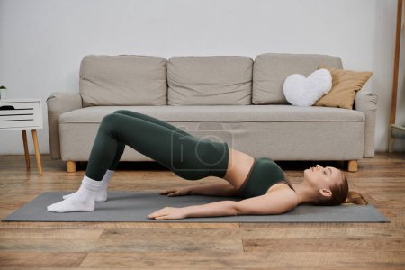 A young woman in green workout clothes performs a bridge pose on a yoga mat in her living room.