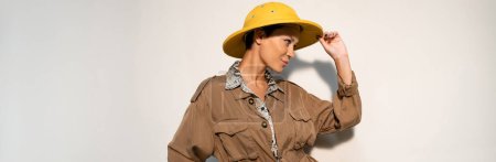 young archaeologist touching yellow safari hat and looking away on grey background, banner