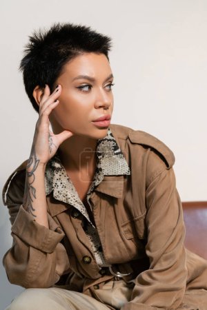 Photo for Brunette archaeologist in beige jacket with tattooed hand near face looking away on grey - Royalty Free Image