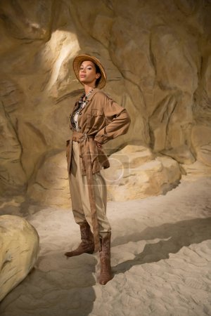 Foto de Longitud completa de arqueólogo en sombrero de safari y ropa beige de pie con la mano en la cadera en la cueva - Imagen libre de derechos