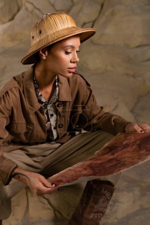 Archäologe im Safari-Outfit betrachtet alte Landkarte, während er in der Höhle sitzt