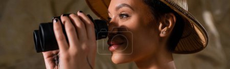 young archaeologist in safari hat looking through binoculars in desert, banner