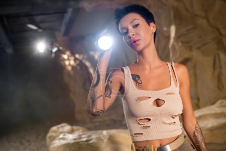 junger tätowierter Archäologe im Tanktop hält Taschenlampe in der Hand und blickt in Höhle in die Kamera
