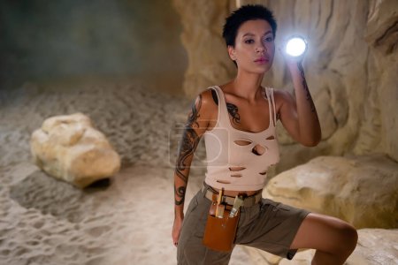 Photo for Sexy archaeologist in tank top and waist bag with brushes holding flashlight in cave - Royalty Free Image