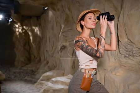 Foto de Sexy y tatuado arqueólogo en safari sombrero sosteniendo binoculares y mirando a la cámara en el desierto - Imagen libre de derechos