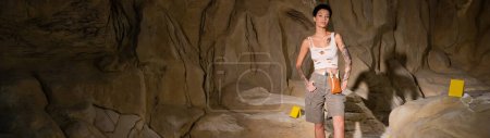 sexy tattooed archaeologist standing with hands on hips near rock in cave, banner