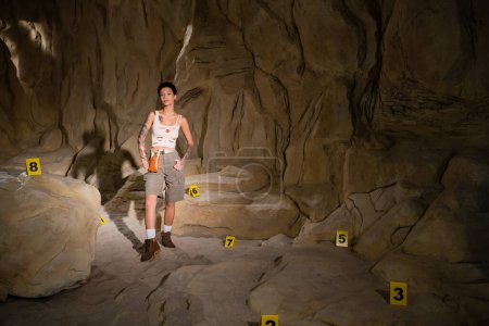 tattooed archaeologist in shorts and tank top standing near numbered marks in cave