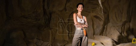 joven arqueólogo morena en pantalones cortos y camiseta de pie con los brazos cruzados en la cueva oscura, pancarta