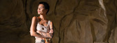 tattooed archaeologist in sexy tank top standing with crossed arms and looking away in cave, banner Poster #617058136
