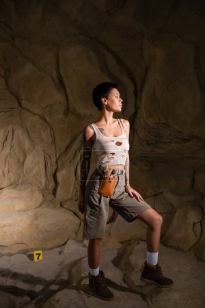 full length of tattooed brunette archaeologist in tank top and shorts looking away in dark cave