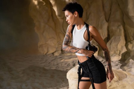 sexy brunette archaeologist in crop top and shorts touching holster with gun in desert
