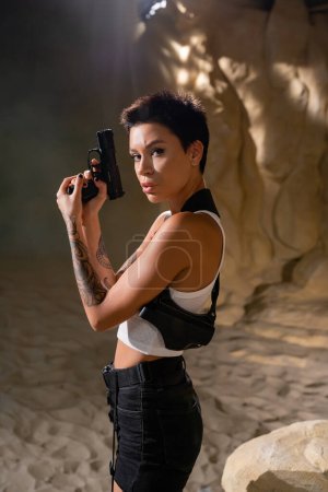 brunette archéologue avec étui tenant pistolet et regardant caméra dans le désert