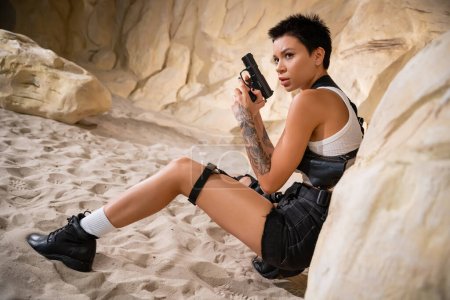 Sexy Archäologin mit kurzen Haaren und Tätowierung hält Waffe in der Hand, während sie sich hinter Felsen in Höhle versteckt