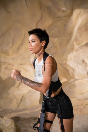 Foto de Arqueólogo armado y tatuado en traje sexy corriendo en cueva - Imagen libre de derechos