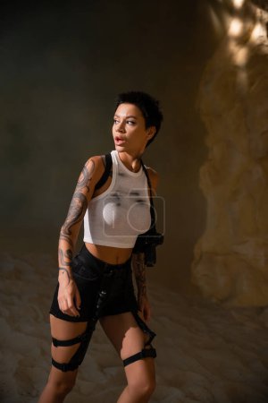 curioso y tatuado arqueólogo en traje sexy con pistola en funda caminando en cueva