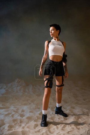 full length of sexy and armed archaeologist with tattoo and dirty crop top looking away while standing on sand