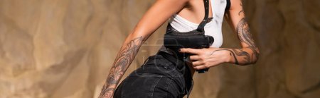 Photo for Cropped view of dangerous and tattooed archaeologist with holster holding gun, banner - Royalty Free Image