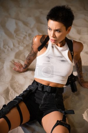 vista de ángulo alto de arqueólogo tatuado y sexy con funda y pistola sentado en la arena 