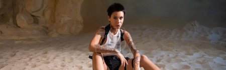 tattooed and sexy archaeologist in dirty crop top and shorts sitting on sand, banner