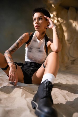 sexy young archaeologist with tattoo and short hair sitting in dirty clothes on sand