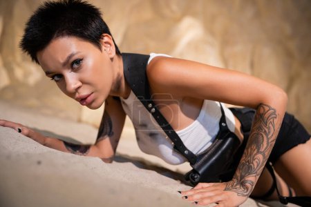 sexy y tatuado arqueólogo en ropa sucia y funda arrastrándose sobre la arena en la cueva