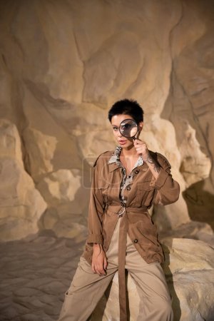 Junge Archäologin mit Tätowierung hält Lupe in der Nähe des Gesichts in Höhle