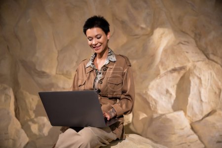 arqueólogo feliz con tatuaje y pelo corto usando el ordenador portátil en la cueva durante la expedición