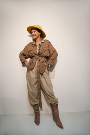 Téléchargez les photos : Pleine longueur de heureux archéologue en chapeau safari et veste beige posant avec les mains dans les poches sur gris - en image libre de droit