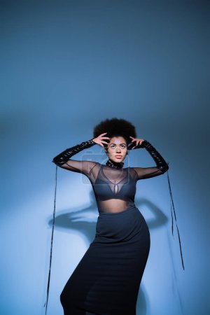 stylish african american woman in transparent shirt and black bra looking up on blue