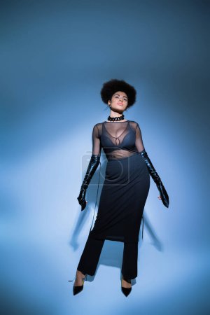 full length of curly african american woman in transparent shirt and black skirt looking up on blue