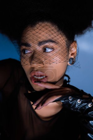 portrait of curly african american woman in black veil looking away while touching lip on blue 
