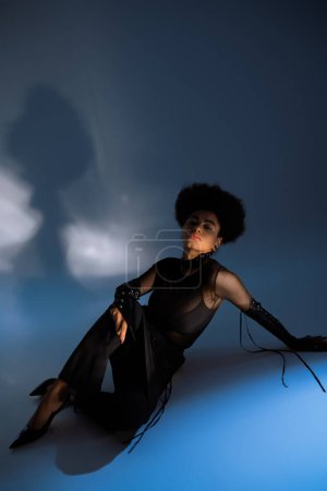 Foto de Mujer americana africana rizada en camisa transparente y falda negra sentada en azul - Imagen libre de derechos