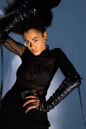 trendy african american model in black outfit posing with hand on hip on blue 