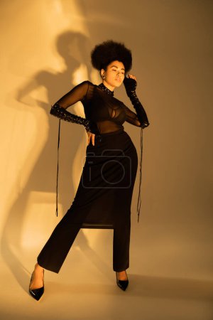 volle Länge des lockigen afrikanisch-amerikanischen Modells in sexy transparentem Outfit posiert mit der Hand an der Hüfte auf gelb 