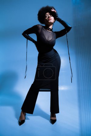 in voller Länge von lockigem afrikanisch-amerikanischem Model im schwarzen Outfit posiert mit der Hand an der Hüfte auf blau