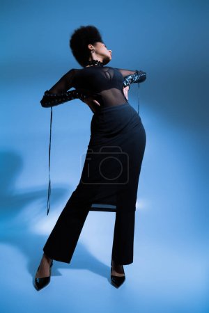 full length of curly african american model in black outfit posing with hands on waist on blue