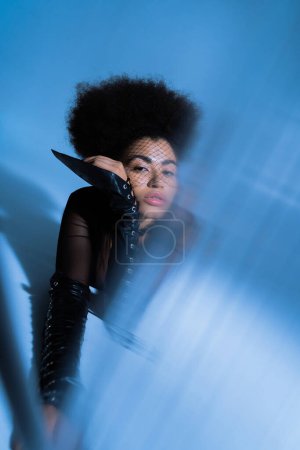 Foto de Joven mujer afroamericana en velo negro mirando a la cámara mientras posa en azul con el primer plano borroso - Imagen libre de derechos
