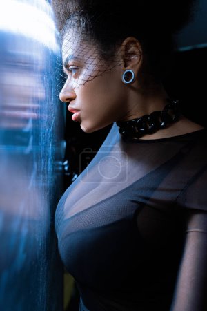 young african american woman in sexy outfit and black veil looking through glass with blue lighting 