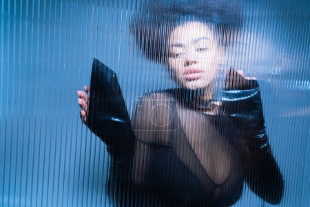 joli modèle afro-américain en tenue sexy noire regardant loin à travers le verre froissé