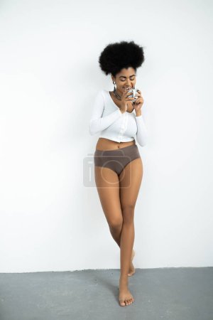 full length of happy african american woman in long sleeve shirt and panties holding cup of coffee on white 
