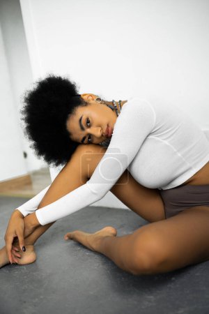 barefoot african american woman in long sleeve shirt and panties sitting on grey floor near white wall