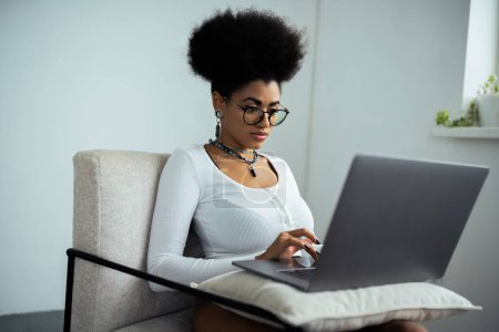 concentrated african american woman in eyeglasses using laptop while sitting on armchair 