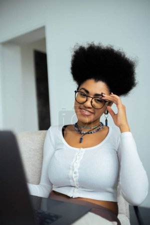 happy african american freelancer in eyeglasses smiling near laptop at home