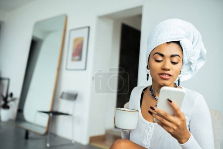 hübsche Afroamerikanerin mit Handtuch auf dem Kopf, Smartphone und Kaffeetasse in der Hand 